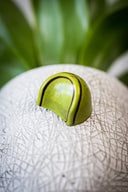 A single lime green bon bon sits atop a white sphere with plant life in the background. 