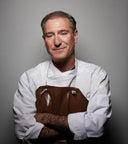 A portrait of a man wearing a white chef's coat and a brown leather apron, standing with arms crossed against a plain gray studio background.
