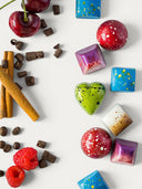 Norman Love Confections assorted dark chocolates, including heart-shaped, spherical, and square pieces in vibrant colors like red, green, blue, and purple, with garnishes of cherries, raspberries, cinnamon sticks, and coffee beans.