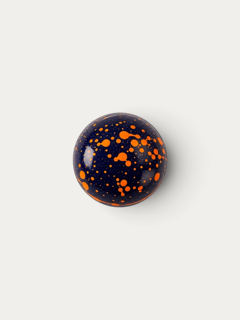 Norman Love Confections Vanilla Caramel chocolate sphere with a glossy dark blue shell and bright orange splatter